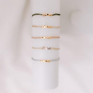 May include: Five gold bracelets with the word "oma" on them. The bracelets are on a white background and are stacked on top of each other. The bracelets are made of different colored cords, including green, beige, and gray.