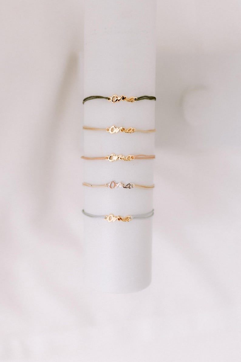 May include: Five gold bracelets with the word "oma" on them. The bracelets are on a white background and are stacked on top of each other. The bracelets are made of different colored cords, including green, beige, and gray.