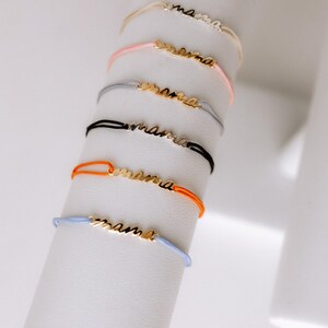May include: A set of six colourful string bracelets with a gold 'mama' charm. The bracelets are displayed on a white stand.