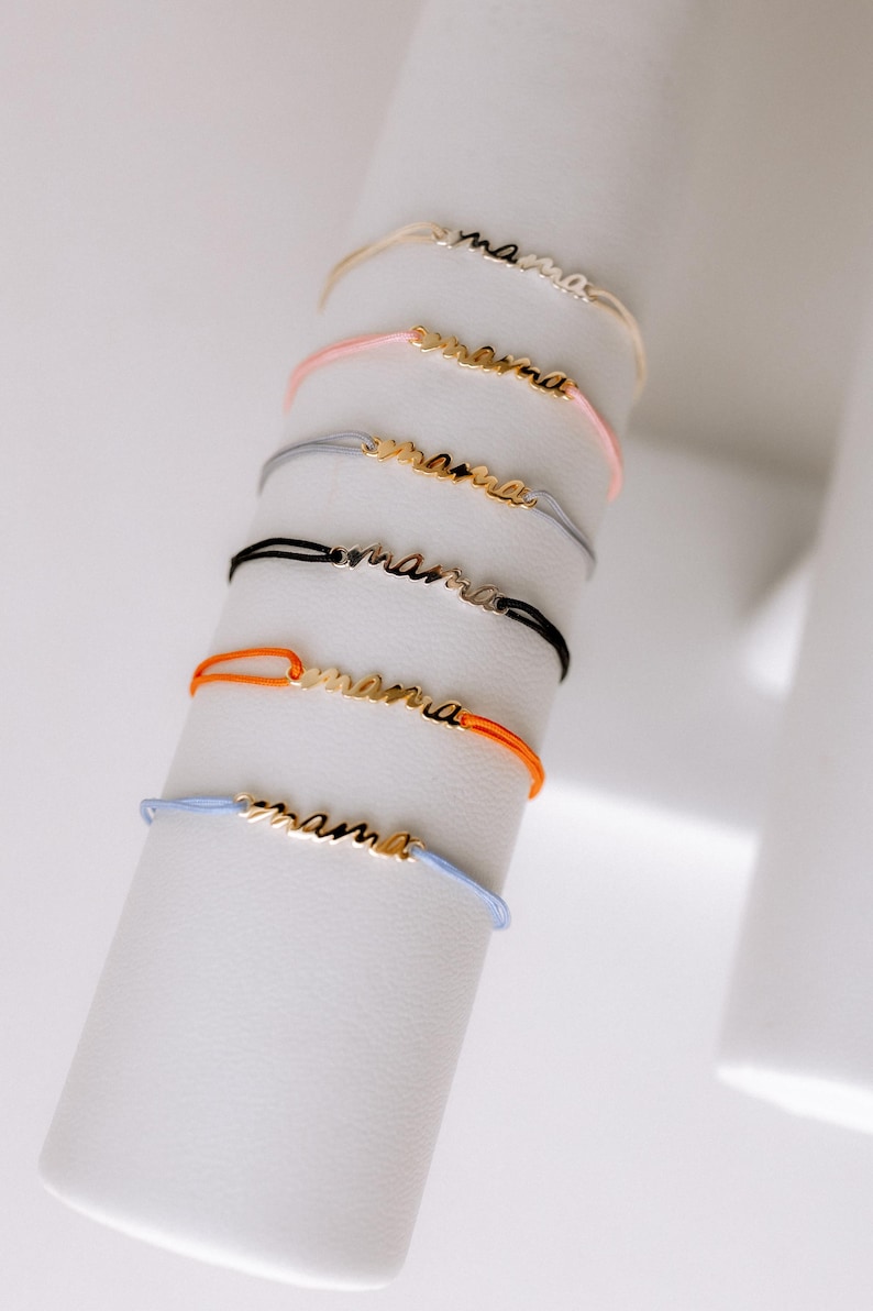 May include: A set of six colourful string bracelets with a gold 'mama' charm. The bracelets are displayed on a white stand.