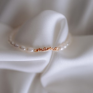 May include: A delicate gold necklace with the word "mama" spelled out in cursive script. The necklace is made of small white pearls.