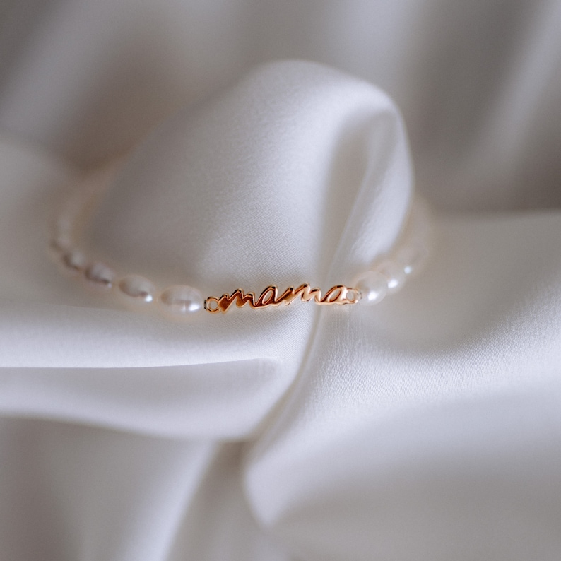 May include: A delicate gold necklace with the word "mama" spelled out in cursive script. The necklace is made of small white pearls.