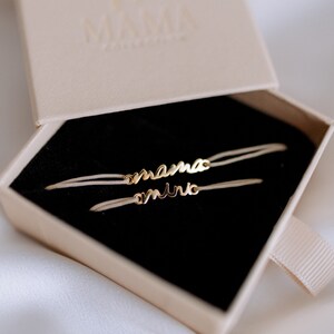 May include: Two delicate bracelets with the words "mama" and "mini" in gold lettering on a white cord. The bracelets are displayed in a black velvet box with a beige lid.