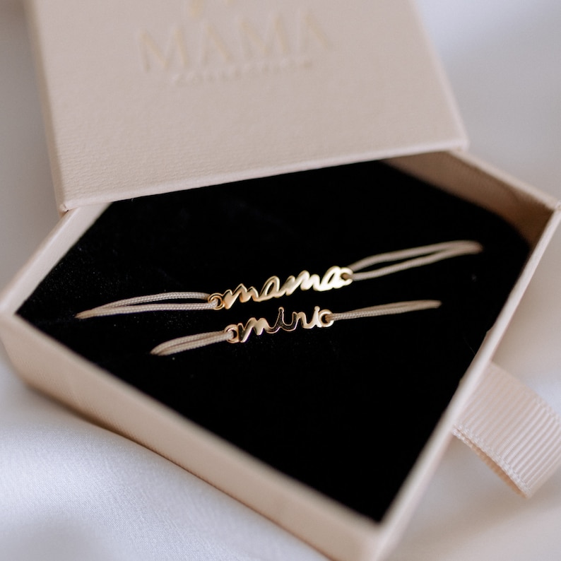 May include: Two delicate bracelets with the words "mama" and "mini" in gold lettering on a white cord. The bracelets are displayed in a black velvet box with a beige lid.