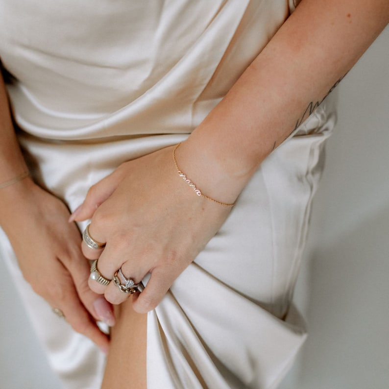May include: A gold chain bracelet with the word "mama" spelled out in cursive. The bracelet is on a person's wrist, and they are wearing several rings on their fingers.