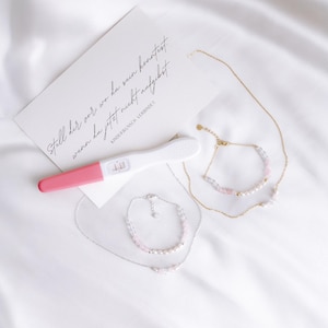 Puede incluir: Una prueba de embarazo rosa y blanca con un resultado positivo, junto con dos delicados collares con cuentas rosas y blancas. El texto en la tarjeta dice "Stell dir vor wo du sun konntest wenn du jetzt nicht aufgegeben" y "Kinderwunsch verbindet".