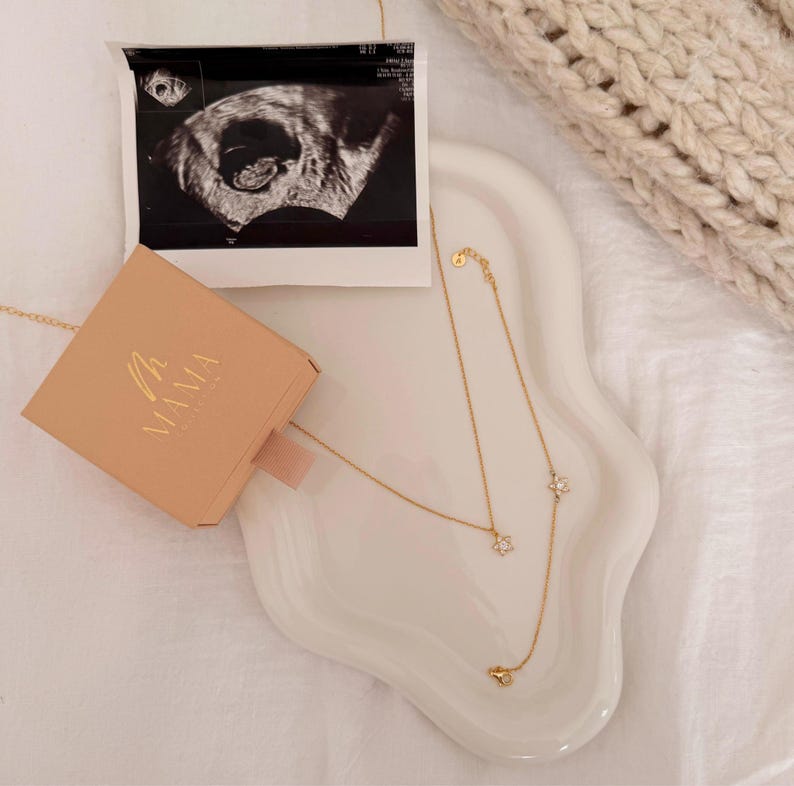 May include: A gold necklace with flower pendants and a matching bracelet are displayed on a white, wavy tray. A small, peach-colored box with the word "MAMA" in gold lettering sits next to an ultrasound image.