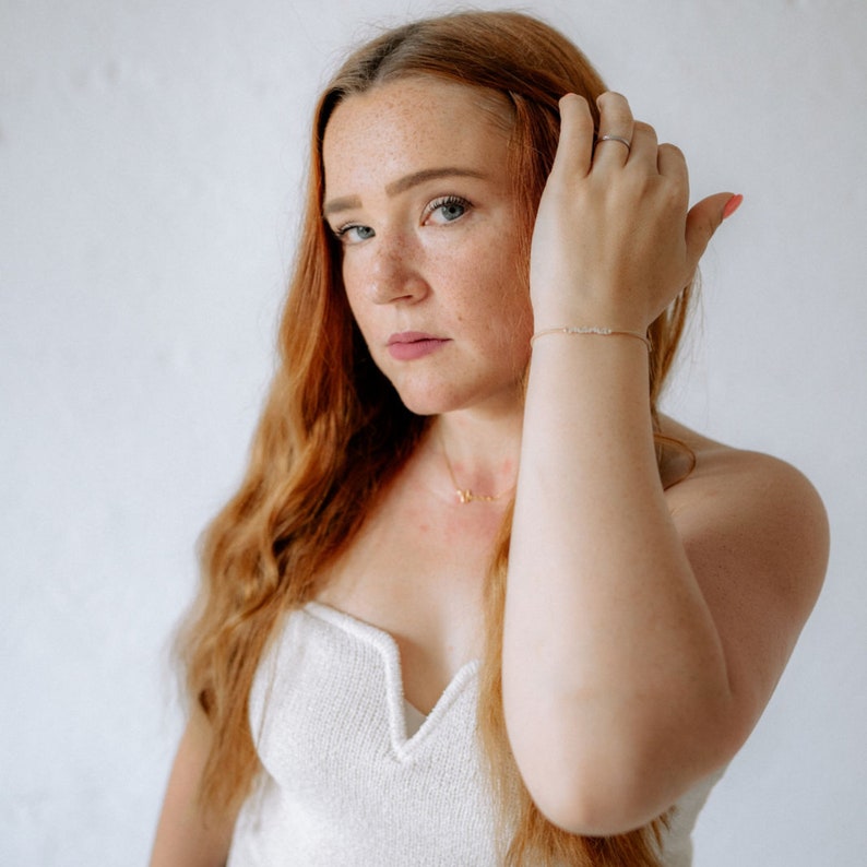 May include: A woman with long, wavy red hair is wearing a white, sleeveless top. She is looking at the camera with a serious expression. She is wearing a gold necklace and a gold bracelet with small, clear stones.