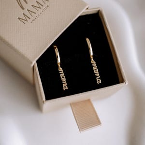 May include: A pair of gold hoop earrings with the word "mama" dangling from each hoop. The earrings are in a small, beige box with the words "The Mama Collection" printed on the top.