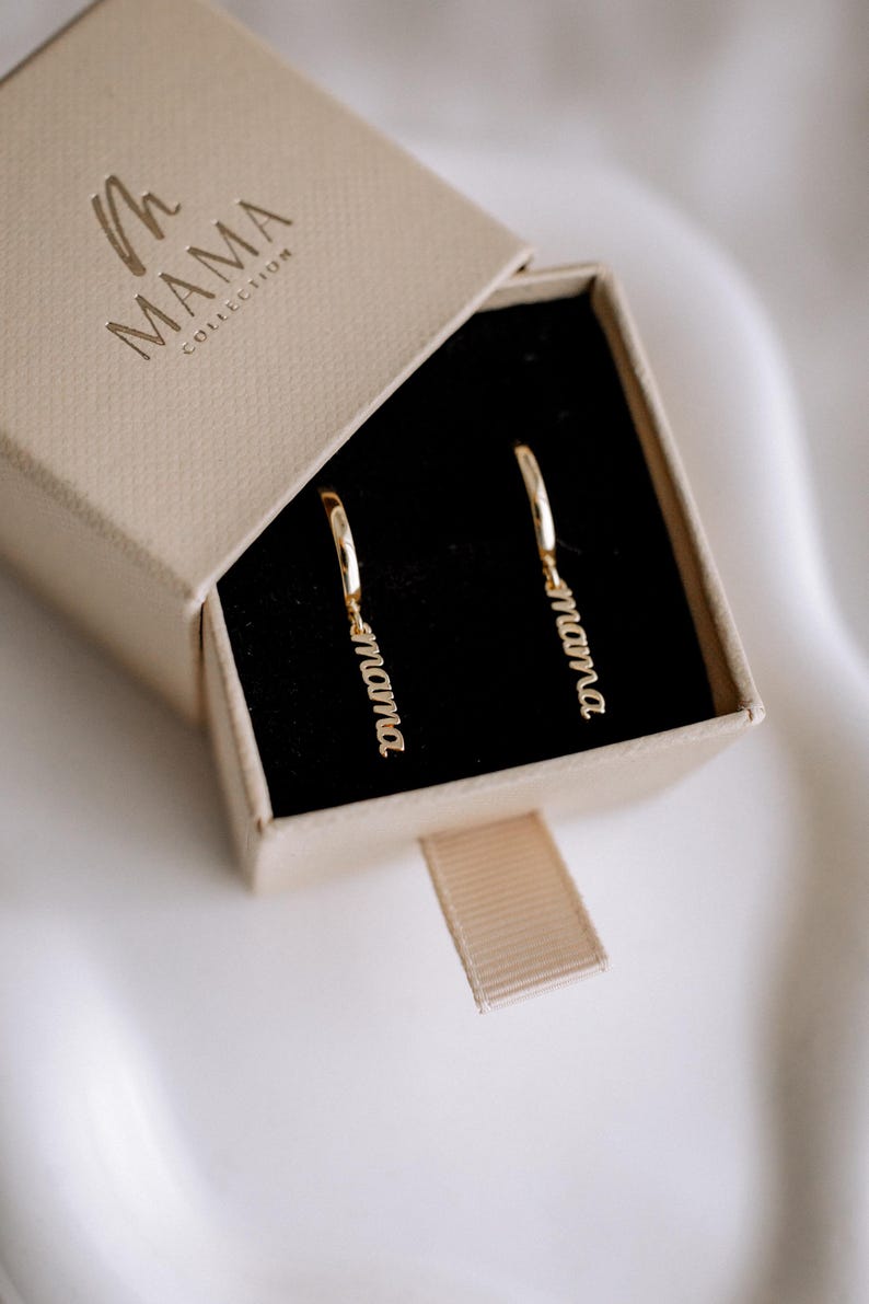 May include: A pair of gold hoop earrings with the word "mama" dangling from each hoop. The earrings are in a small, beige box with the words "The Mama Collection" printed on the top.