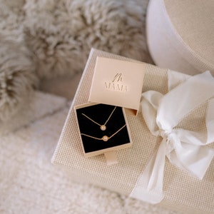 May include: A white gift box with a beige ribbon and a smaller box inside. The smaller box is open and contains two gold necklaces with small round pendants. The smaller box has the text "MAMA" on the lid.