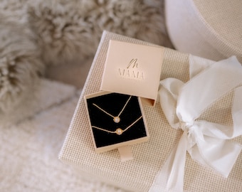 May include: A white gift box with a beige ribbon and a smaller box inside. The smaller box is open and contains two gold necklaces with small round pendants. The smaller box has the text "MAMA" on the lid.