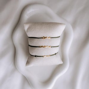 May include: Three delicate gold bracelets with the words "oma", "mama", and "mini" on a white cushion. The bracelets are on a white background.