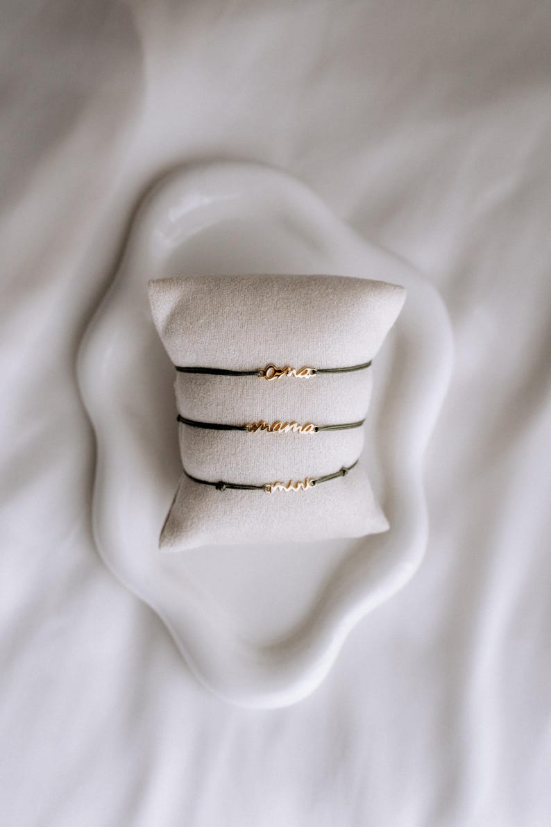 May include: Three delicate gold bracelets with the words "oma", "mama", and "mini" on a white cushion. The bracelets are on a white background.