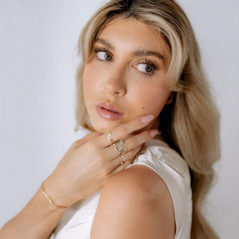 May include: A woman wearing a white top with a gold bracelet and three gold rings on her finger. The rings are decorated with small diamonds. The woman is wearing a gold ring with the word "LOVE" spelled out in diamonds.