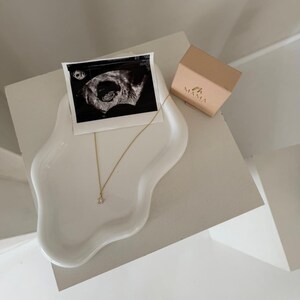 May include: A white, organically shaped ceramic tray holds a black and white ultrasound image, a gold necklace with a small pendant, and a small, tan box with the word "MAMA" printed on it. The items are arranged on a white surface.