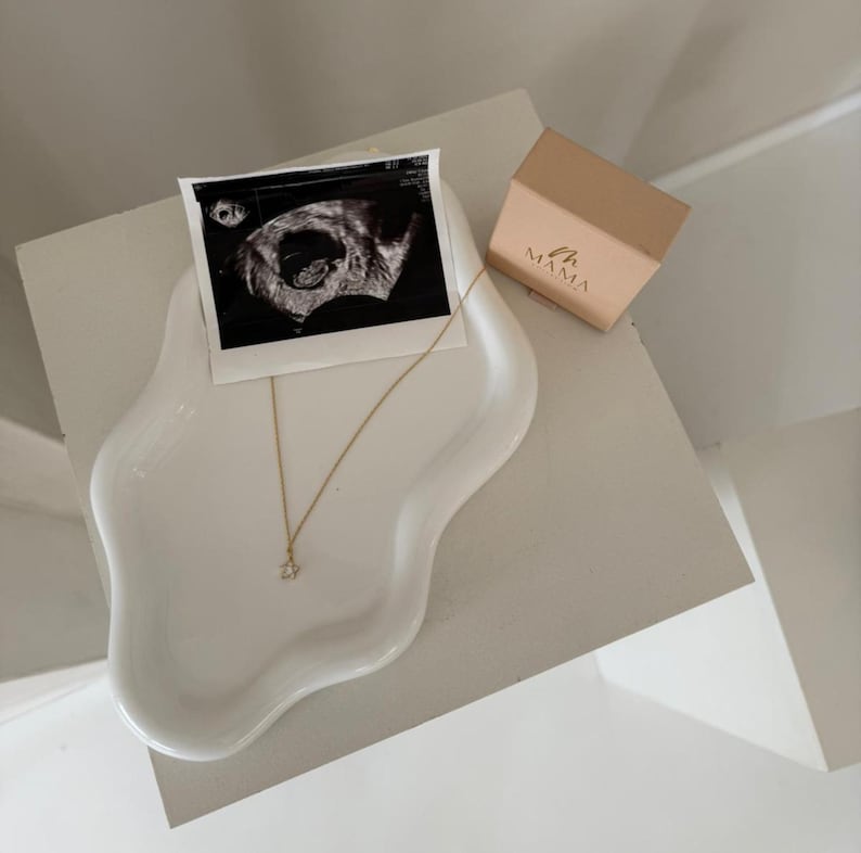May include: A white, organically shaped ceramic tray holds a black and white ultrasound image, a gold necklace with a small pendant, and a small, tan box with the word "MAMA" printed on it. The items are arranged on a white surface.