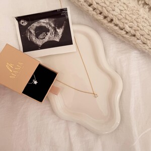 May include: A gold necklace with a star pendant rests on a white wavy tray, next to a small box with the word "MAMA" on it. An ultrasound image is also on the tray. The necklace is on a white surface.