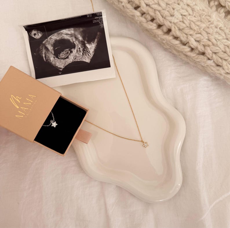 May include: A gold necklace with a star pendant rests on a white wavy tray, next to a small box with the word "MAMA" on it. An ultrasound image is also on the tray. The necklace is on a white surface.