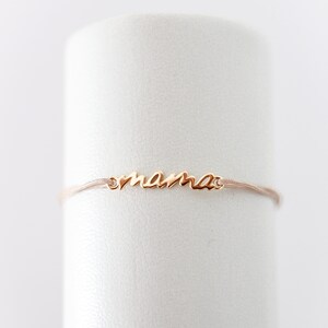May include: A delicate gold bracelet with the word "mama" inscribed on it. The bracelet is made of a thin, light brown cord.