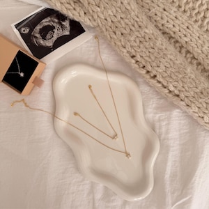 May include: A collection of delicate gold necklaces with star pendants displayed on a white, wavy-edged ceramic tray. A small gift box with a star necklace is open. An ultrasound image and a beige knit sweater are in the background.