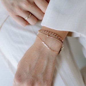 May include: A woman's wrist with three gold bracelets. The bottom bracelet has the word "mama" spelled out in cursive. The middle bracelet has a small round charm. The top bracelet is a thick chain.