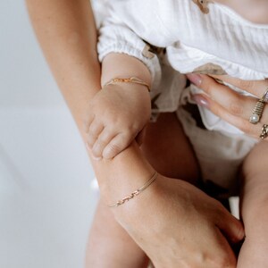 May include: A close-up of two wrists wearing delicate gold bracelets. The bracelets are simple and elegant, with a thin chain and small charms. The bracelets are worn on the wrists of a mother and child, symbolising a loving bond.