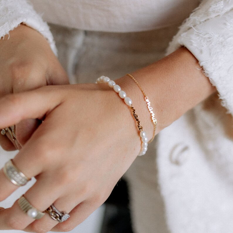 May include: A gold chain bracelet with the word "mama" on it, layered over a pearl bracelet with the word "remarkable" on it.