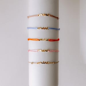 May include: Five bracelets with the word "amitie" in gold lettering on a white background. The bracelets are in different colours: brown, blue, orange, pink, and beige.