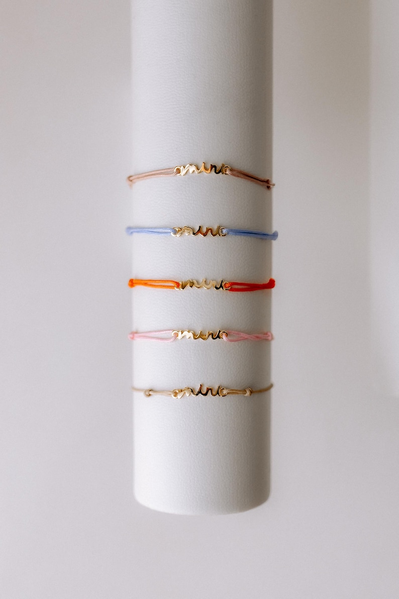 May include: Five bracelets with the word "amitie" in gold lettering on a white background. The bracelets are in different colours: brown, blue, orange, pink, and beige.