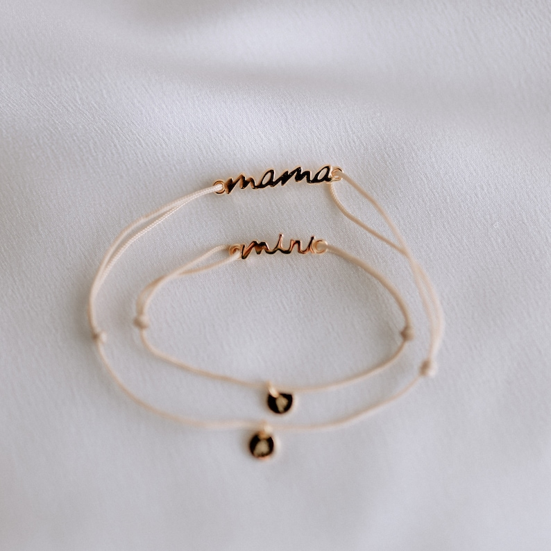 May include: Two delicate bracelets with white string and gold charms. The larger bracelet has the word "mama" on it, and the smaller bracelet has the word "mini" on it.