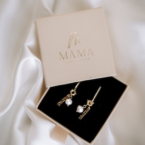 Pu&ograve; includere: Un paio di orecchini d'oro con un ciondolo di perla e la parola "DAD" e "MOM" appesi a ciascun orecchino. Gli orecchini sono in una piccola scatola beige con il logo "MAMA COLLECTION" stampato sulla parte superiore.