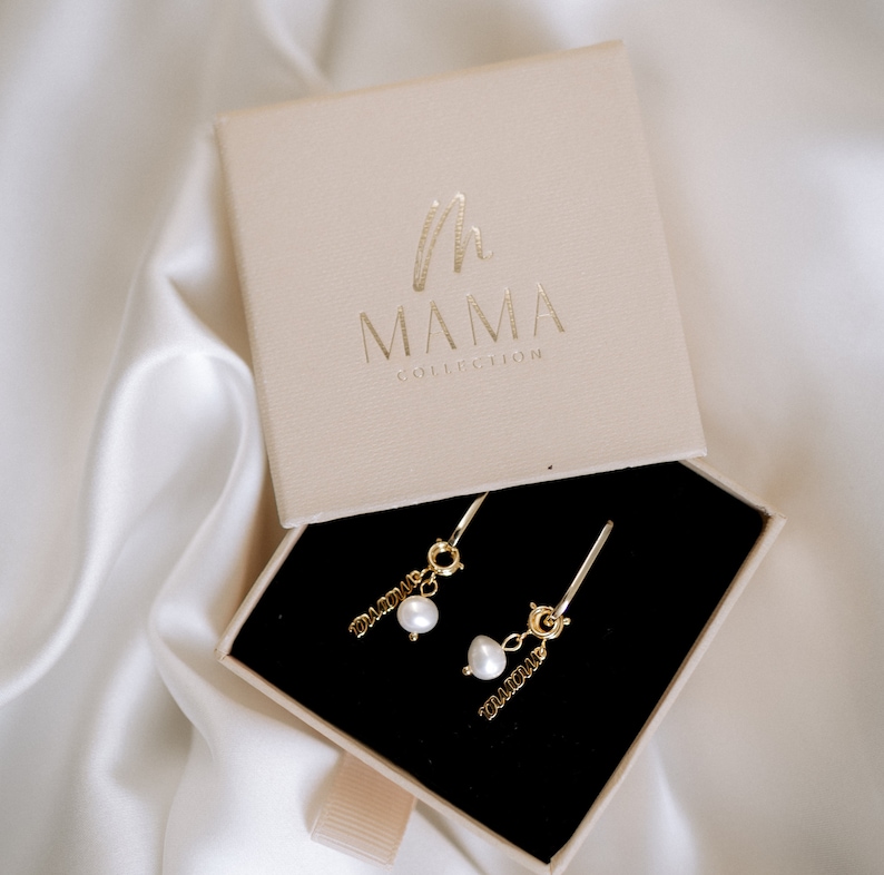 Pu&ograve; includere: Un paio di orecchini d'oro con un ciondolo di perla e la parola "DAD" e "MOM" appesi a ciascun orecchino. Gli orecchini sono in una piccola scatola beige con il logo "MAMA COLLECTION" stampato sulla parte superiore.