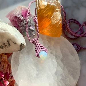 May include: A pink macrame necklace with a white opal pendant. The necklace is displayed on a white selenite plate with other crystals.