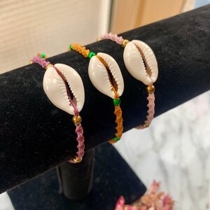 May include: Three bracelets featuring a white seashell charm, each bracelet is made with a different coloured cord: pink, orange, and brown. The cords are accented with small green and gold beads.