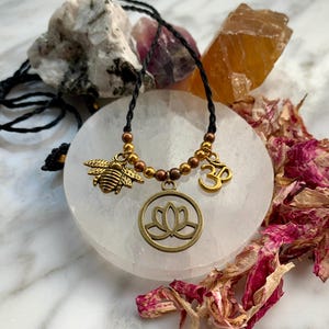 Puede incluir: Un collar de cordón negro con cuentas doradas y dijes. Los dijes incluyen una abeja, un símbolo Om y una flor de loto. El collar se muestra sobre una superficie blanca.