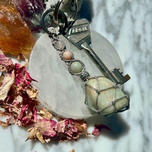 Peut inclure: Un porte-clés composé d'une clé en métal argenté avec le texte ARCU gravé sur la tête. Un cordon en macramé gris clair retient trois petites perles rondes rose pâle et vertes, se terminant par une pierre lisse vert clair enveloppée dans un filet.