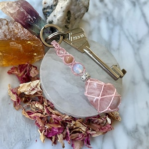 Puede incluir: Un llavero con una llave de metal plateado unida a un cordón de macramé rosa claro. El cordón está tejido con una pequeña cuenta redonda color melocotón, una cuenta translúcida tipo ópalo y una cuenta espaciadora plateada, terminando en cuarzo rosa.