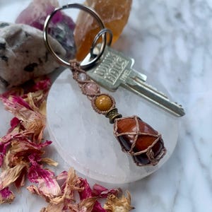 Puede incluir: Una piedra de ojo de tigre marrón y roja envuelta en macramé con un llavero adjunto. La piedra está rodeada de pequeñas cuentas en tonos marrones, rosas y amarillos.