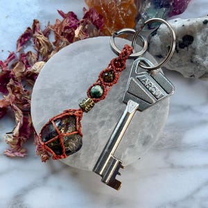 May include: A keychain featuring a silver-colored metal key with the word ARCU engraved on the head, attached to a rust-colored macrame cord. The cord is woven with three small round beads and ends in a larger, dark, polished stone encased in netting.