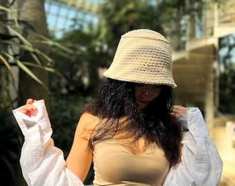 May include: A woman wearing a white crochet bucket hat, a beige tank top, a white shirt, and blue jeans. She is standing in a greenhouse.