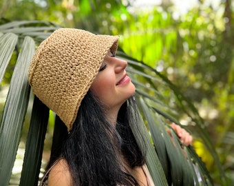 May include: A woman wearing a light brown crochet bucket hat with a wide brim. She is standing in a lush green garden with palm leaves.
