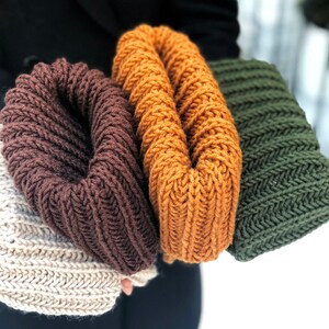 Puede incluir: Tres bufandas infinitas de punto en marr&oacute;n, naranja y verde. Las bufandas est&aacute;n dobladas y se sostienen en la mano de una persona.