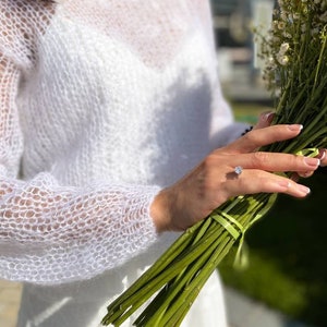 May include: A white, sheer, knitted sweater with long sleeves and a round neckline. The sweater is worn by a person holding a bouquet of white flowers with green stems.