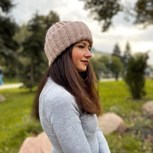 Puede incluir: Una mujer con un gorro de punto de color marr&oacute;n claro y un su&eacute;ter gris. Est&aacute; sentada en una roca en un parque.