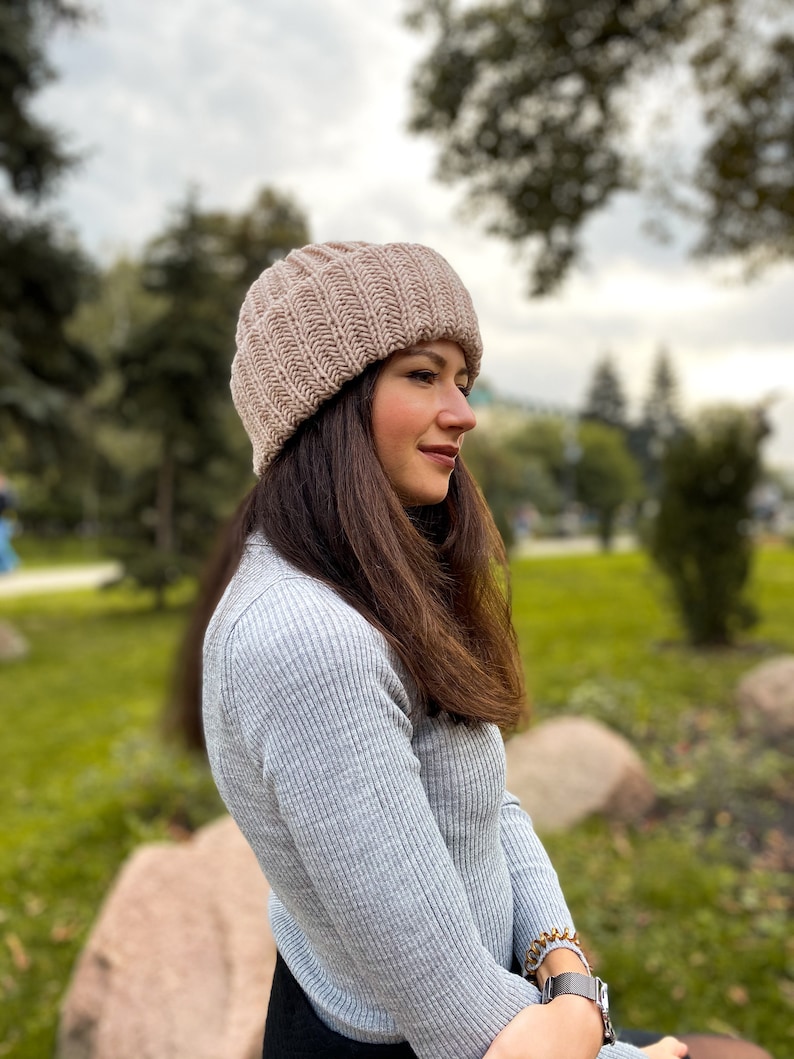 Puede incluir: Una mujer con un gorro de punto de color marr&oacute;n claro y un su&eacute;ter gris. Est&aacute; sentada en una roca en un parque.