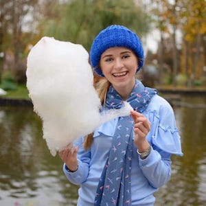 Puede incluir: Una mujer con un gorro azul de punto y una bufanda azul con un patr&oacute;n de p&aacute;jaros blancos sostiene un gran algod&oacute;n de az&uacute;car blanco frente a un estanque.