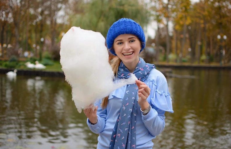 Puede incluir: Una mujer con un gorro azul de punto y una bufanda azul con un patr&oacute;n de p&aacute;jaros blancos sostiene un gran algod&oacute;n de az&uacute;car blanco frente a un estanque.
