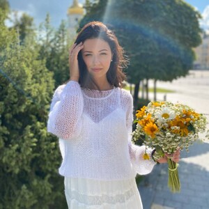 May include: A woman in a white crochet sweater and a white skirt holds a bouquet of yellow and white flowers.