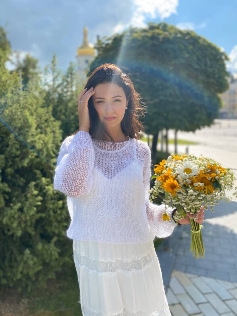 May include: A woman in a white crochet sweater and a white skirt holds a bouquet of yellow and white flowers.